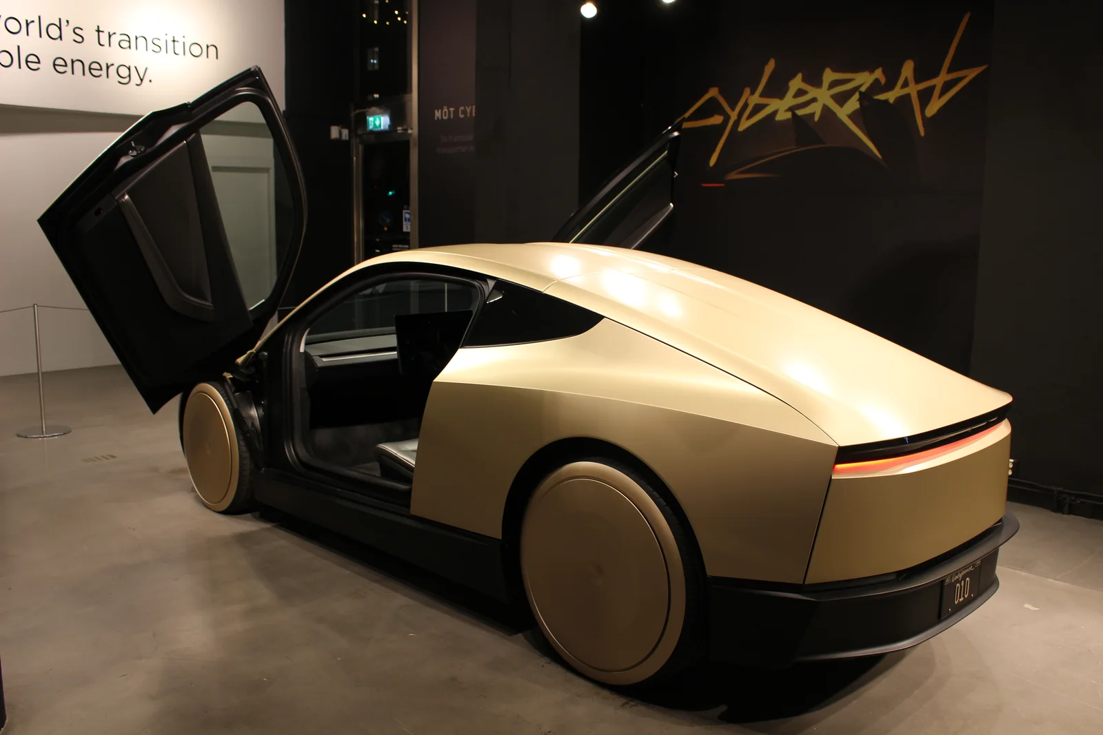 Rear three-quarter view of a gold Tesla Cybercab on display with both doors raised.
