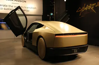 Rear three-quarter view of a gold Tesla Cybercab on an indoor display, with both doors raised.